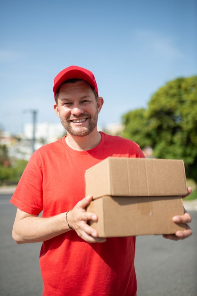 Happy deliveryman in a red shirt and cap holding packages outdoors on a sunny day.